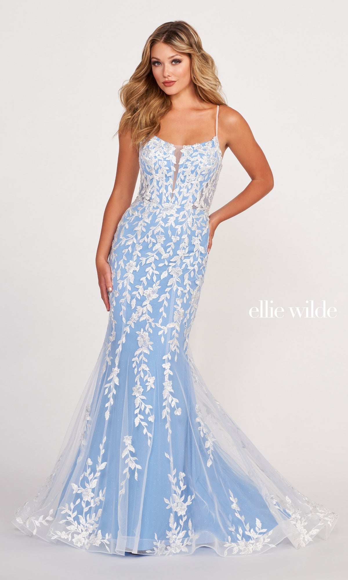 A woman models the Lace Applique Over Glitter Jersey Gown EW34090, a light blue, floor-length dress with white embroidered lace and thin straps, posing with one hand on her hip.